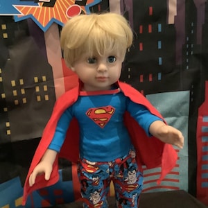 Puede incluir: Una mu&ntilde;eca vestida de Superman, con una camisa azul con el logo de Superman, una capa roja y pantalones estampados. La mu&ntilde;eca tiene el pelo rubio y botas marrones. El fondo presenta un paisaje urbano y la palabra "KAP!"