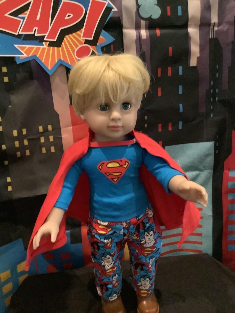 Puede incluir: Una mu&ntilde;eca vestida de Superman, con una camisa azul con el logo de Superman, una capa roja y pantalones estampados. La mu&ntilde;eca tiene el pelo rubio y botas marrones. El fondo presenta un paisaje urbano y la palabra "KAP!"