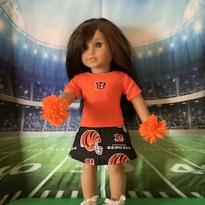 May include: A doll wearing a black and orange cheerleader skirt with the Cincinnati Bengals logo. The doll is also wearing an orange t-shirt with the number 13 and a Bengals logo. The doll is holding orange pom-poms in each hand.