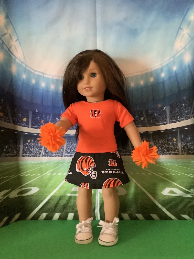 May include: A doll wearing a black and orange cheerleader skirt with the Cincinnati Bengals logo. The doll is also wearing an orange t-shirt with the number 13 and a Bengals logo. The doll is holding orange pom-poms in each hand.