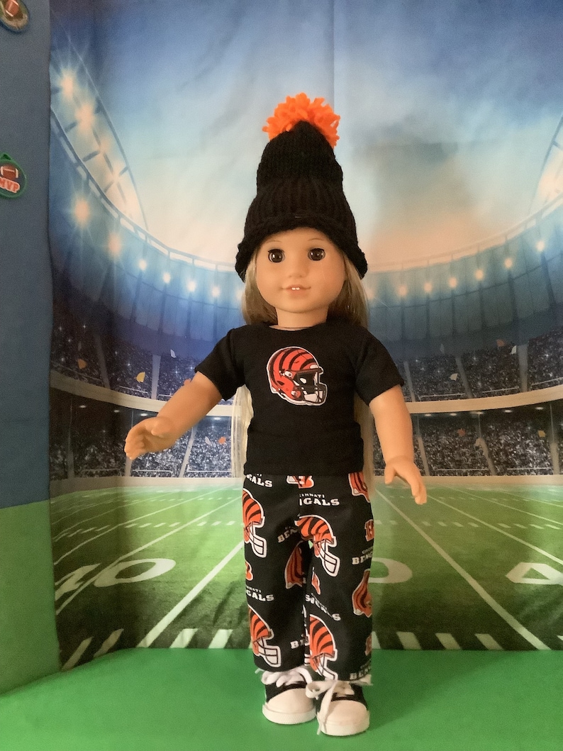 May include: A doll wearing a black and orange beanie with a pom pom, a black t-shirt with a Cincinnati Bengals logo, and black and orange pants with the Cincinnati Bengals logo. The doll is standing on a green and white football field.