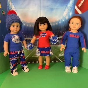 May include: Three dolls dressed in Buffalo Bills football team attire. The dolls are wearing blue and red shirts with the Bills logo, blue pants, and blue skirts. The dolls are holding pom-poms and are standing on a green surface.
