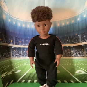 May include: A black and red doll wearing a black jumpsuit with the words "Cincinnati Bengals" on the front. The doll has brown curly hair and brown eyes. The doll is standing on a green surface in front of a stadium backdrop.
