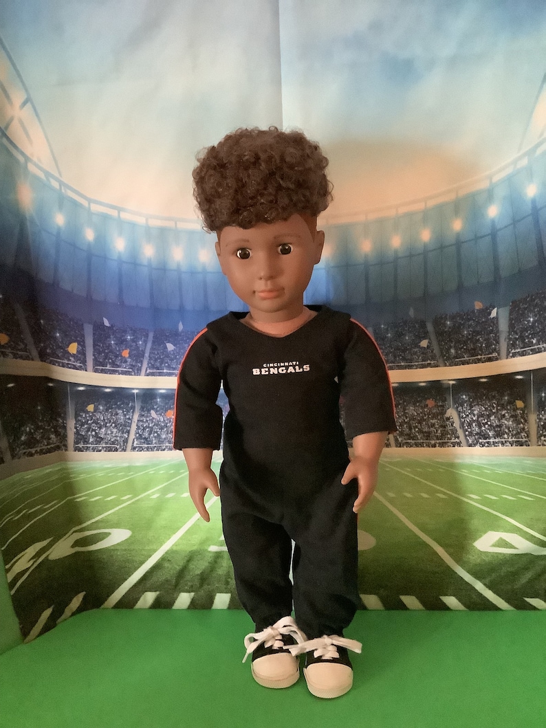 May include: A black and red doll wearing a black jumpsuit with the words "Cincinnati Bengals" on the front. The doll has brown curly hair and brown eyes. The doll is standing on a green surface in front of a stadium backdrop.
