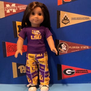 May include: A doll wearing a purple t-shirt with the LSU logo and yellow and purple pajama pants with the LSU logo. The doll is standing in front of a blue background with college pennants.