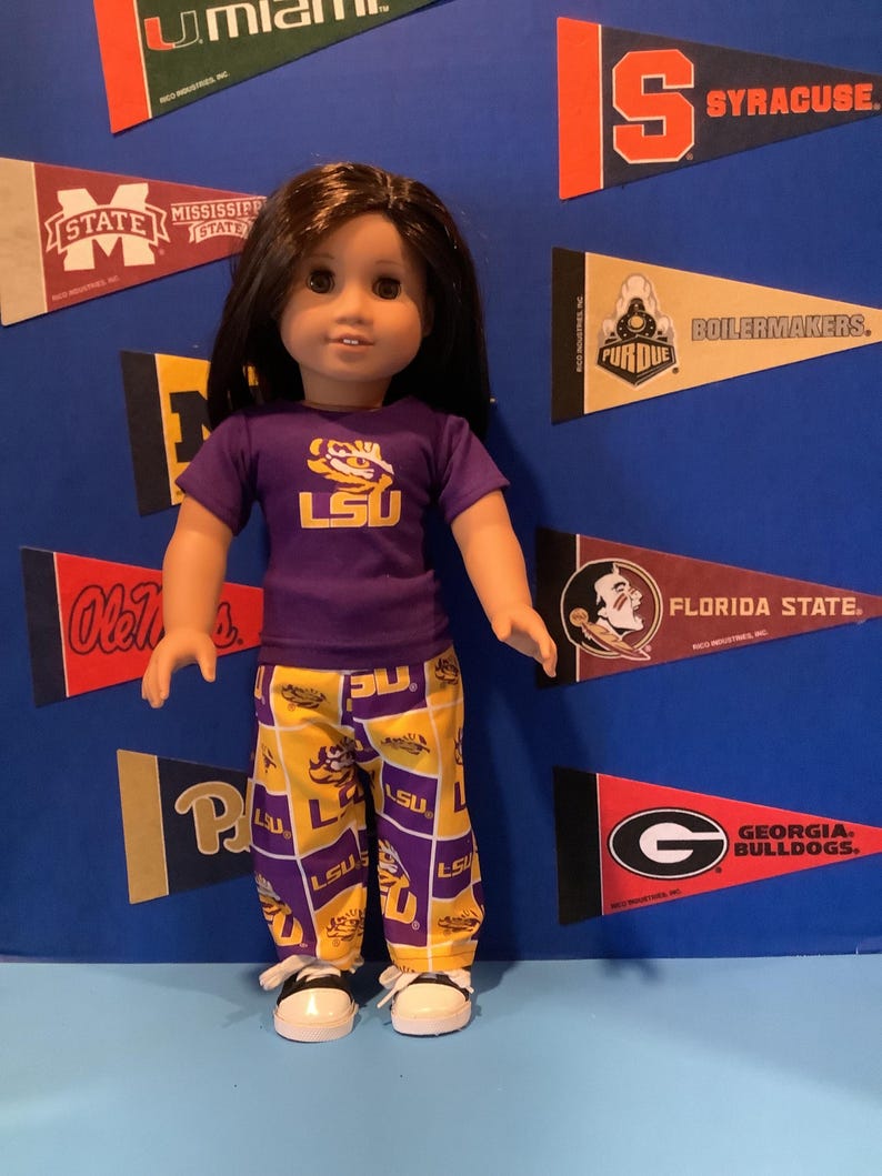May include: A doll wearing a purple t-shirt with the LSU logo and yellow and purple pajama pants with the LSU logo. The doll is standing in front of a blue background with college pennants.