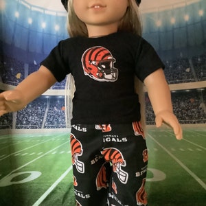 May include: A doll wearing a black t-shirt with a Cincinnati Bengals logo and black and orange pants with a repeated Bengals logo. The doll is wearing a black beanie and has blonde hair.
