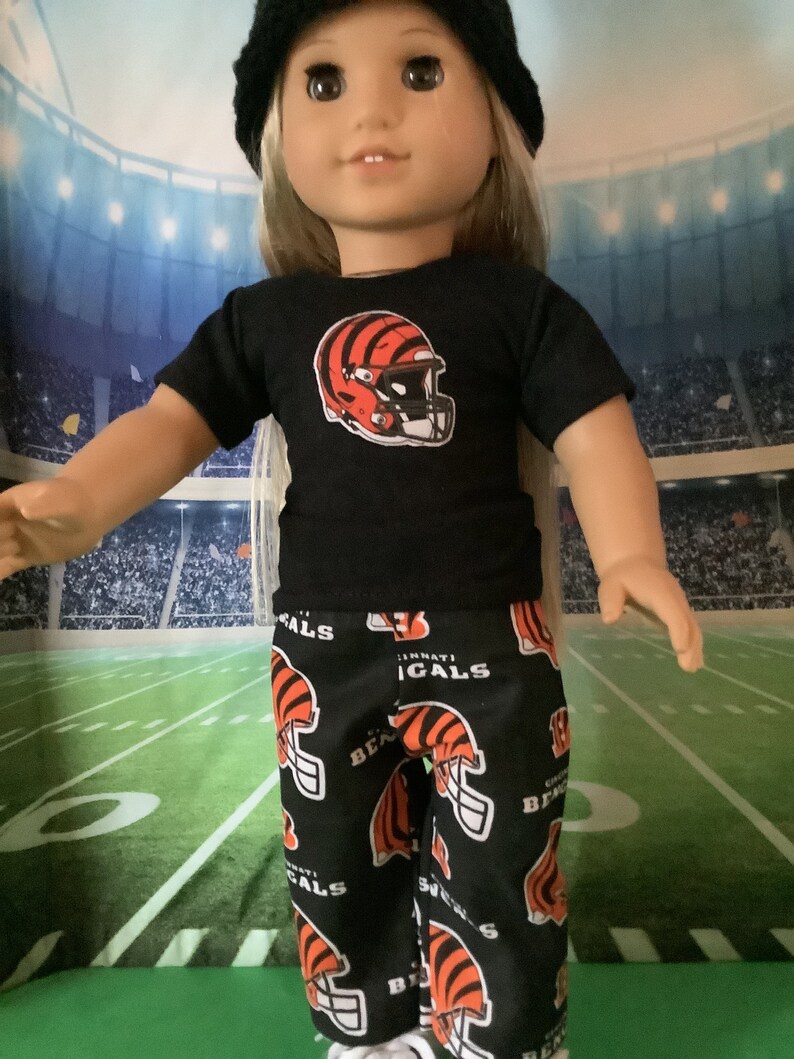 May include: A doll wearing a black t-shirt with a Cincinnati Bengals logo and black and orange pants with a repeated Bengals logo. The doll is wearing a black beanie and has blonde hair.