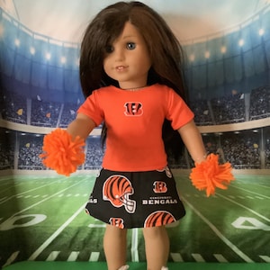 May include: A doll wearing a red and black Cincinnati Bengals outfit with a pom-pom in each hand. The doll is standing in front of a green and white football field.