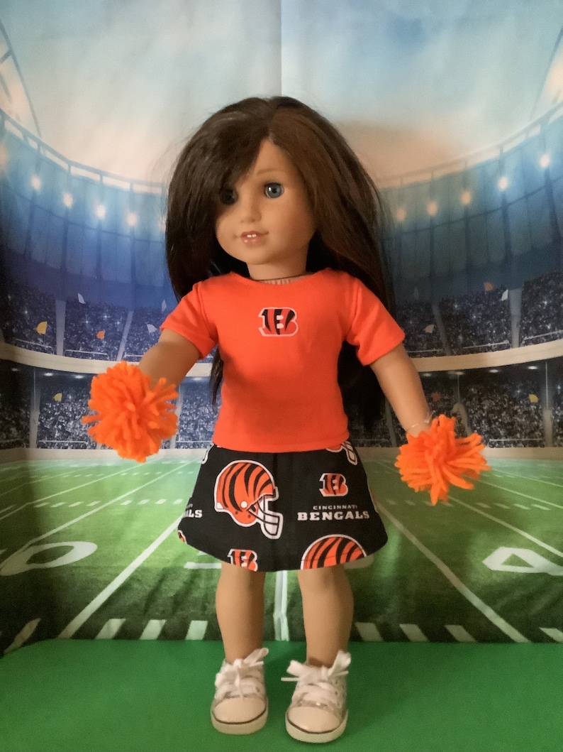 May include: A doll wearing a red and black Cincinnati Bengals outfit with a pom-pom in each hand. The doll is standing in front of a green and white football field.