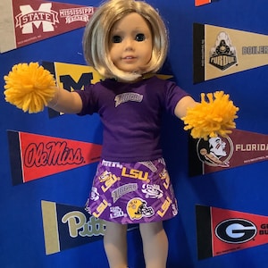 May include: A doll wearing a purple LSU Tigers cheerleading outfit with a yellow pom-pom in each hand. The doll is standing in front of a blue background with college pennants.