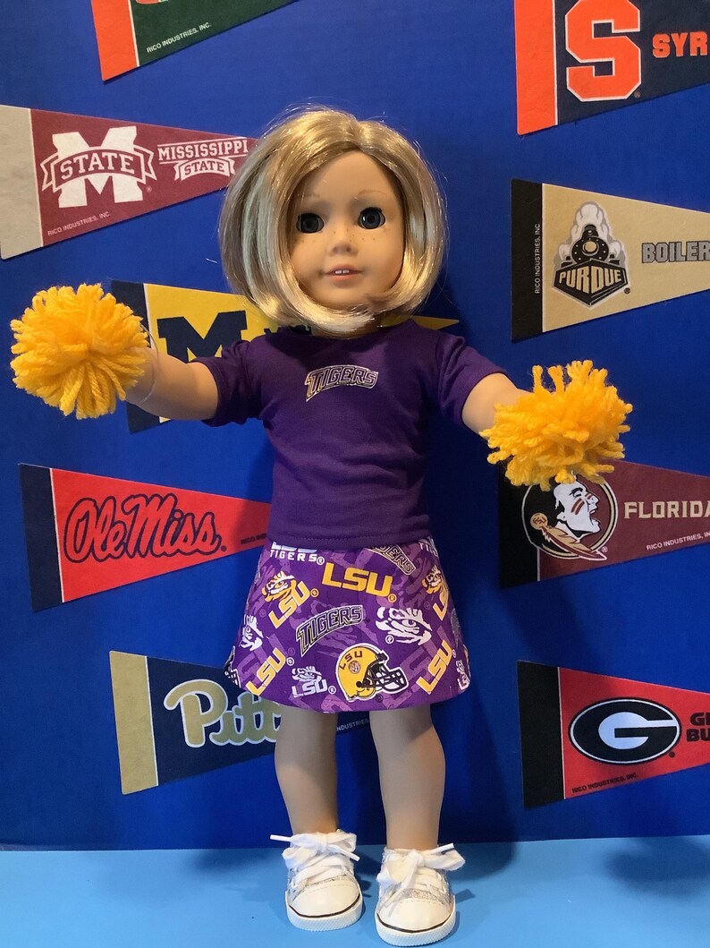 May include: A doll wearing a purple LSU Tigers cheerleading outfit with a yellow pom-pom in each hand. The doll is standing in front of a blue background with college pennants.