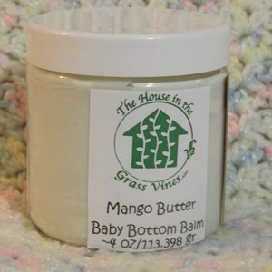 May include: A clear jar with a white lid containing a creamy balm. The label reads "The House in the Grass Vines.us" with a green house logo. Below it says "Mango Butter Baby Bottom Balm" and "~4 OZ/113.398 gr".