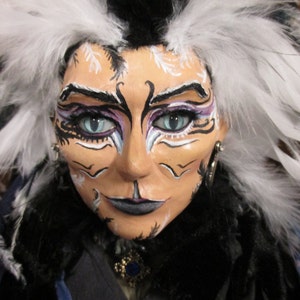 May include: A doll with black and white feathers for hair, wearing a black and blue outfit. The doll has white and black face paint with blue eyes.