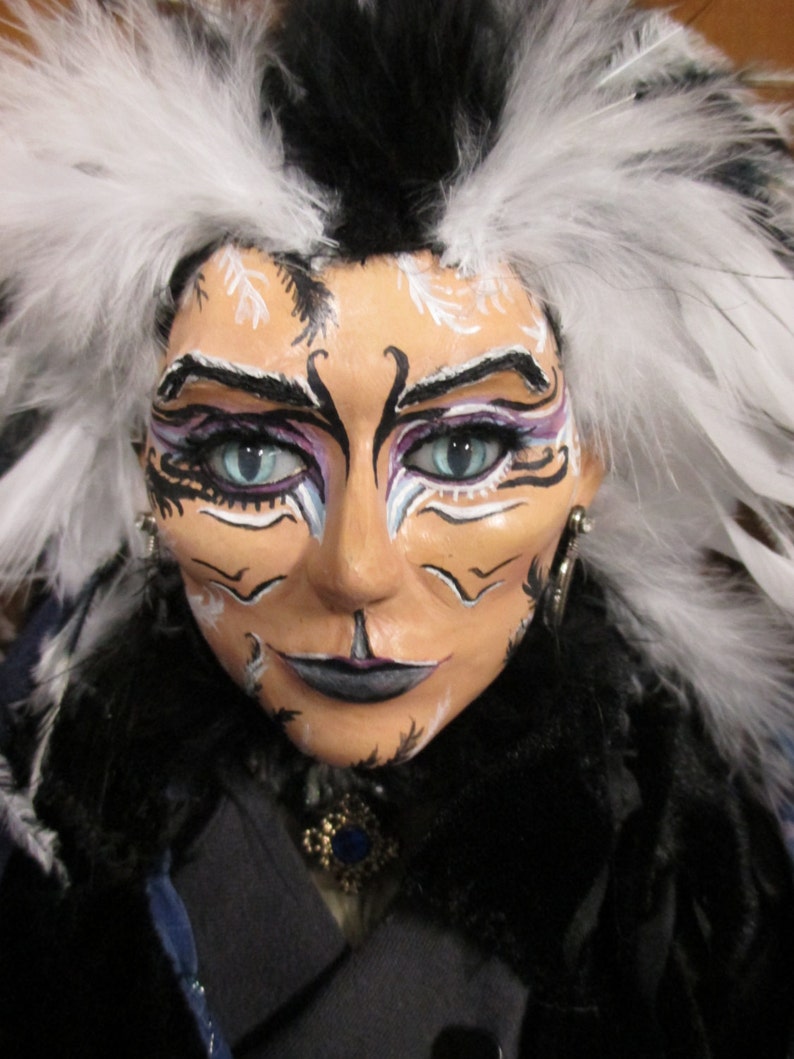 May include: A doll with black and white feathers for hair, wearing a black and blue outfit. The doll has white and black face paint with blue eyes.