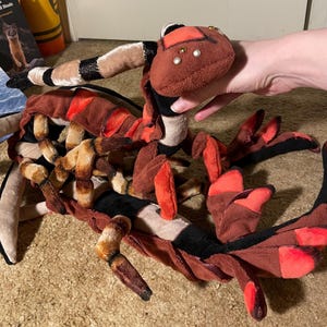 May include: A plush toy scorpion in shades of brown, black, and red. The scorpion has segmented body parts, including legs, a tail, and pincers. The head features small, decorative accents. The toy is designed with a patchwork style.