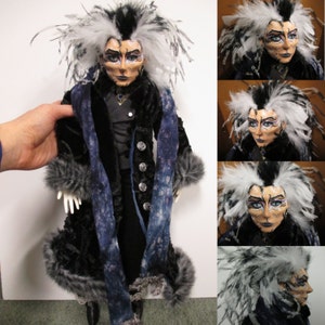 May include: A handmade doll with white and black hair, wearing a black velvet coat with a blue and white scarf. The doll has a painted face with intricate details and is wearing a black velvet coat with a blue and white scarf.