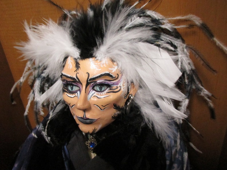 May include: A doll with black and white feathers in its hair. The doll has white face paint with black and purple accents. The doll is wearing a black and white outfit.