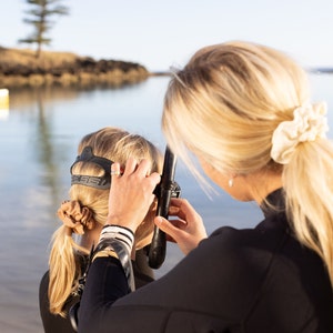 May include: A woman helps another woman put on a black scuba diving mask with a snorkel attached. The woman wearing the mask has brown hair and is wearing a brown hair tie. The woman helping her has blonde hair and is wearing a white hair scrunchie.