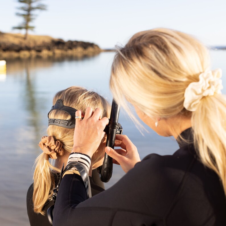 May include: A woman helps another woman put on a black scuba diving mask with a snorkel attached. The woman wearing the mask has brown hair and is wearing a brown hair tie. The woman helping her has blonde hair and is wearing a white hair scrunchie.
