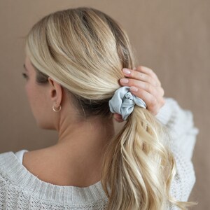May include: A woman with blonde hair is wearing a light blue scrunchie in her hair. She is wearing a light gray sweater.