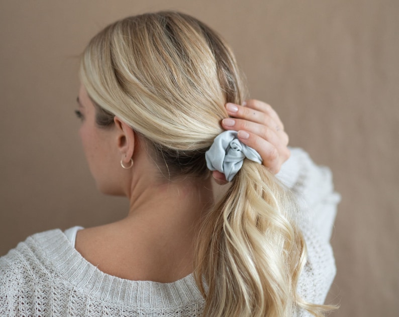 May include: A woman with blonde hair is wearing a light blue scrunchie in her hair. She is wearing a light gray sweater.