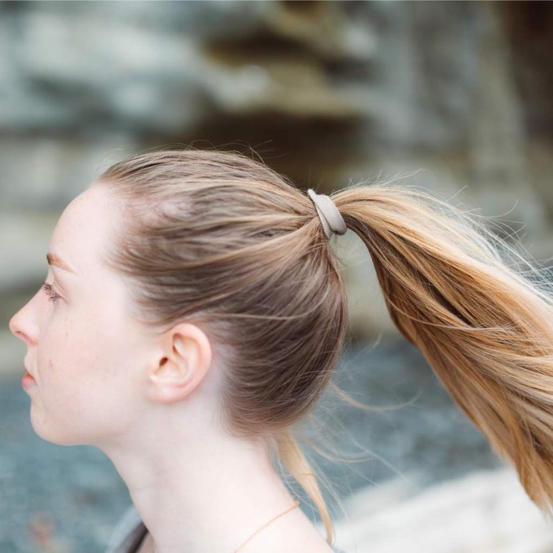May include: A woman with long, light brown hair pulled back in a ponytail with a beige hair tie. Her hair is slightly windblown.