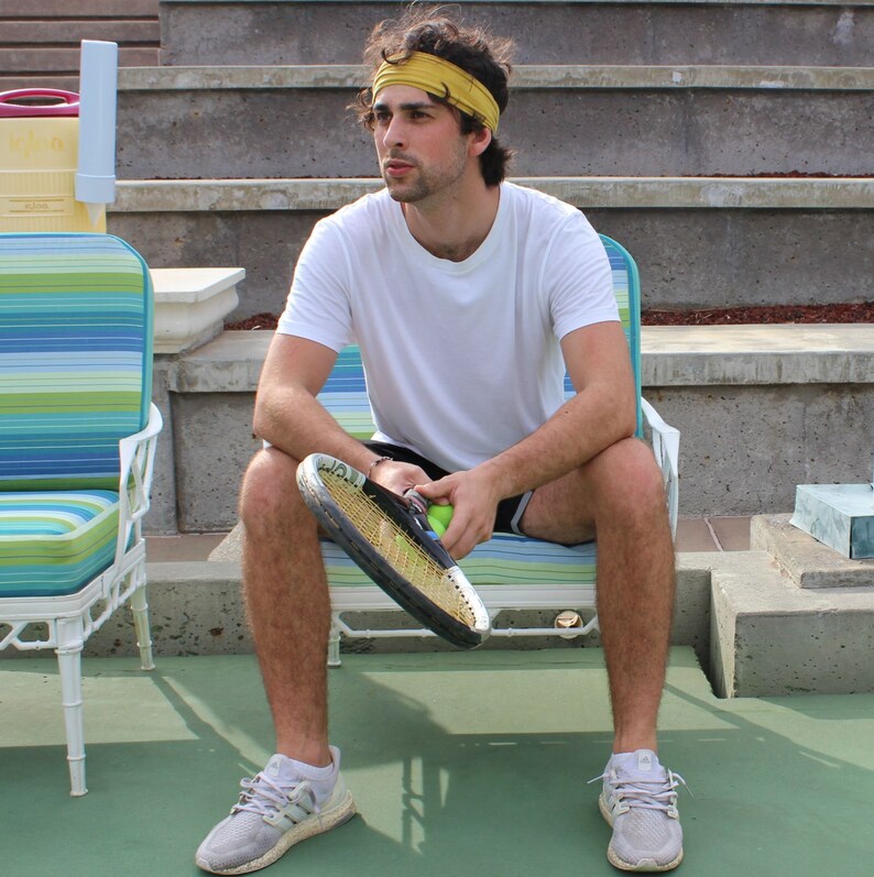May include: A person wearing a yellow headband and white t-shirt sits on a striped chair, holding a tennis racket and ball. The person is wearing grey athletic shoes and black shorts. The background includes concrete steps and a green tennis court.