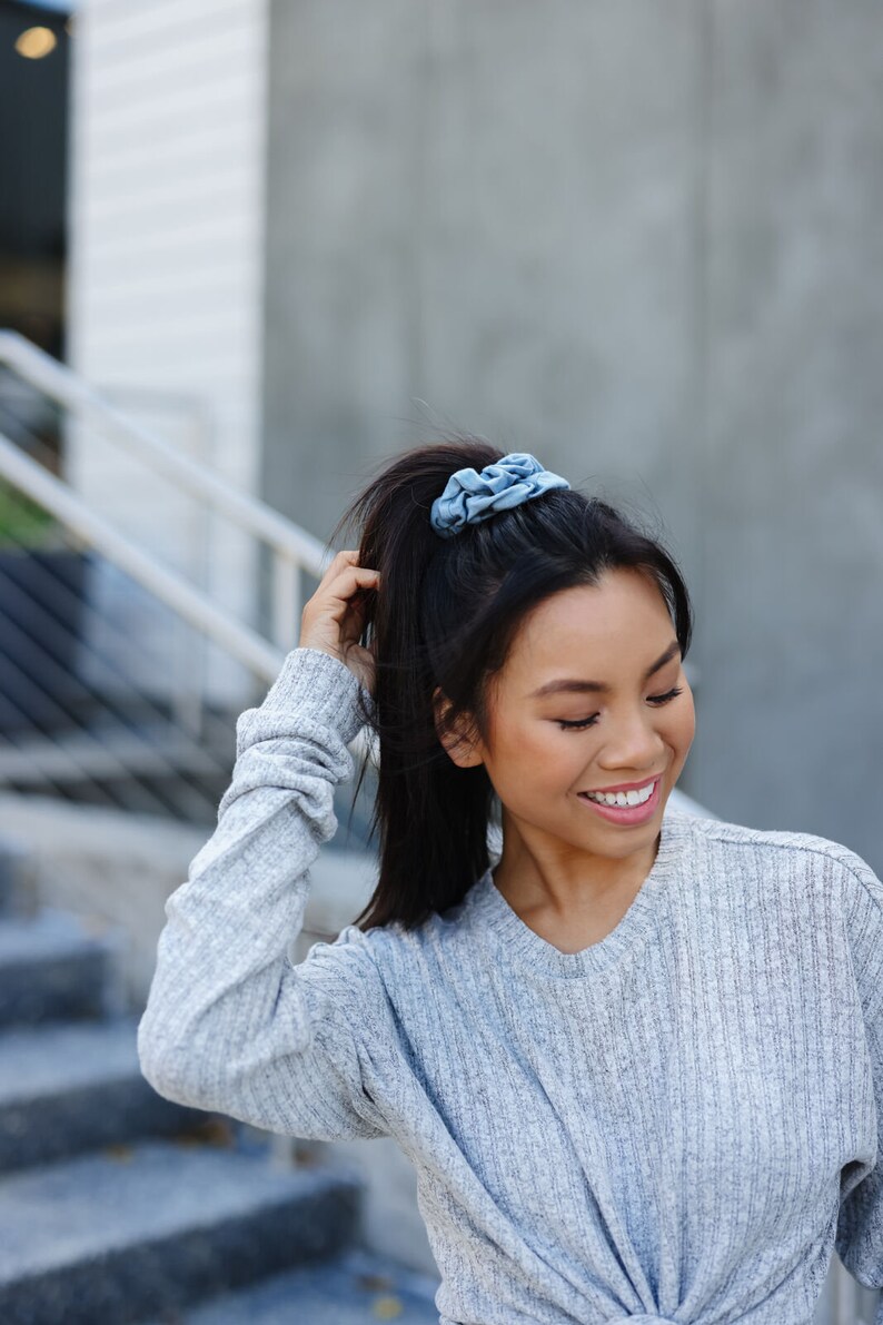 May include: A woman with long dark hair tied up in a ponytail with a blue scrunchie, wearing a light gray sweater.