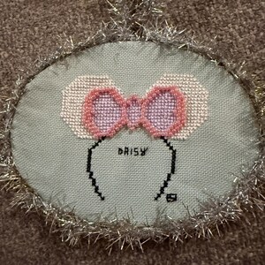 May include: Hand-stitched oval ornament with a light blue background, featuring a cross-stitched pink bow and white ears. The word "DAISY" is stitched below. The ornament is trimmed with silver tinsel.