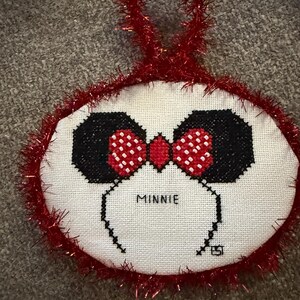 May include: A handmade cross-stitch ornament featuring Minnie Mouse. The ornament has a white background with black ears, a red bow with white polka dots, and the name "MINNIE" stitched below. The edges are trimmed with red tinsel.