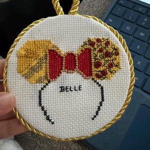May include: A cross-stitched circular ornament with a red bow, gold bead accents, and yellow ears, with the name "BELLE". The ornament is framed with gold rope and has a gold rope loop for hanging.