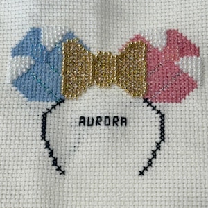 May include: Cross-stitch design on white fabric depicting a bow-shaped headband. The bow has blue, gold, and pink sections. The word "AURORA" is stitched in black below the bow.