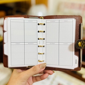 May include: A brown leather six-ring planner with a gold-tone metal binder mechanism. The open planner displays white grid-paper inserts with printed headers for Monday through Sunday and a tab labeled HOME on the bottom right corner.