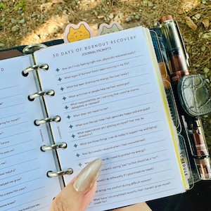 May include: A ring-bound planner page titled 30 Days of Burnout Recovery Journal Prompts, featuring a list of reflective questions marked with star icons. A fountain pen is clipped to the side, and a cat-shaped tab is visible at the top.