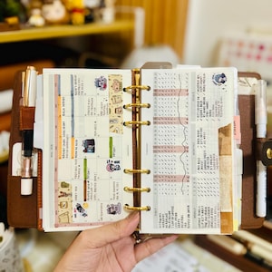 May include: A brown leather six-ring planner open to a monthly calendar and a habit tracker page. The planner features gold-tone rings, a pen holder with a metallic pen, and a second pen holder with a white pen. The pages contain handwritten notes, stickers, and grid-style tracking charts.