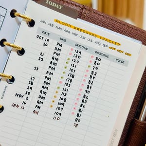 May include: A white paper blood pressure tracker insert for a binder with gold rings. The page features columns for date, time, systolic, diastolic, and pulse readings, with handwritten entries from October to January and small colored dot stickers.