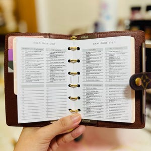 May include: Open brown leather planner with gold rings and a gratitude list. The planner is open to a page with the heading "Gratitude List" and several bulleted lists. A pen is visible on the right side.