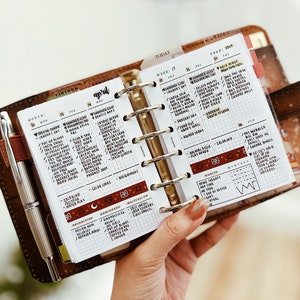 Könnte beinhalten: Ein Nahaufnahme eines braunen Lederplaners mit einer weißen, karierten Seite, die für April geöffnet ist. Der Planer hat goldene Ringe und eine Lasche mit der Aufschrift "Heute". Die Seite ist mit handschriftlichen Notizen und To-Do-Listen gefüllt.