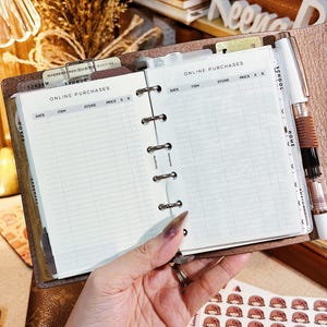 May include: Open brown leather planner with lined pages labeled "ONLINE PURCHASES." A white pen is clipped to the side. The planner is open, revealing a section for tracking online purchases. Decorative stickers are visible.