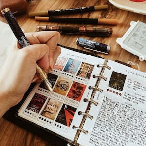 Peut inclure: Un stylo-plume bleu foncé avec un motif de nuit étoilée et des accents dorés est utilisé pour remplir un carnet de lecture. La page présente une grille de couvertures de livres, des notes en étoiles et des commentaires manuscrits sur Verity.