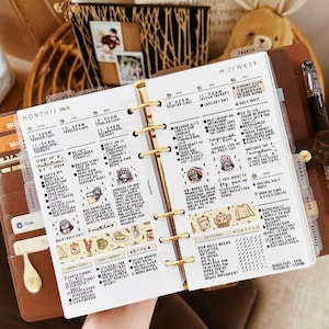 Könnte beinhalten: Ein brauner Lederplaner mit goldenen Akzenten, der einen detaillierten Wochenplan zeigt. Die Seiten sind mit handschriftlichen Notizen, Aufklebern und farbigem Washi-Tape gefüllt, das tägliche Aufgaben und Termine organisiert. Ein Stift und anderes Zubehör sind sichtbar.
