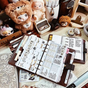 May include: A brown leather ring-bound planner open to a weekly spread with handwritten notes and stickers, surrounded by a plush bear doll, a plush rabbit doll, a white pen holder with markers, a small ink bottle, and a black cutting mat.