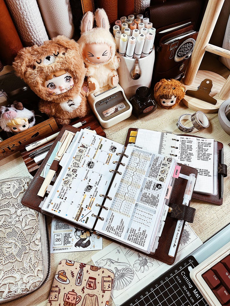 May include: A brown leather ring-bound planner open to a weekly spread with handwritten notes and stickers, surrounded by a plush bear doll, a plush rabbit doll, a white pen holder with markers, a small ink bottle, and a black cutting mat.