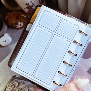 May include: A personal planner insert with a dark brown leather cover, featuring white pages with a grid layout divided into sections labeled GOAL and TASKS. The pages are held together by gold-colored metal rings and include a vertical date tracker.