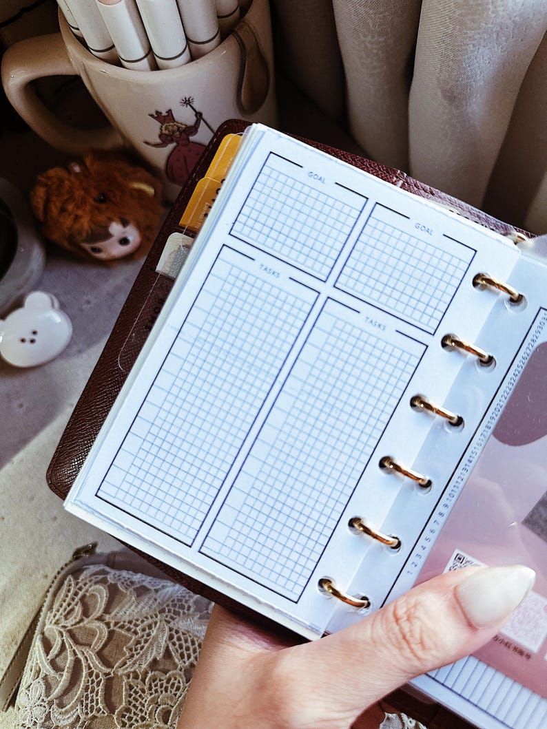 May include: A personal planner insert with a dark brown leather cover, featuring white pages with a grid layout divided into sections labeled GOAL and TASKS. The pages are held together by gold-colored metal rings and include a vertical date tracker.