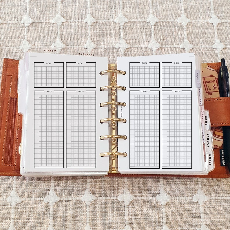 May include: A brown leather ring-bound planner open to a two-page spread with white paper inserts. The pages feature black grid layouts labeled GOAL and TASKS. A gold-colored six-ring mechanism holds the pages. Tabbed dividers labeled AUGUST, SEPTEMBER, OCTOBER, HOME, SCHOOL, and NOTES are visible on the right side, along with a black pen.
