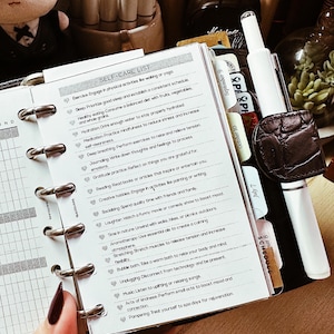 May include: A planner with a self-care list, including items like exercise, healthy eating, and meditation. The planner is open to a page with the title "SELF-CARE LIST". A white pen and a brown leather pen holder are also visible.