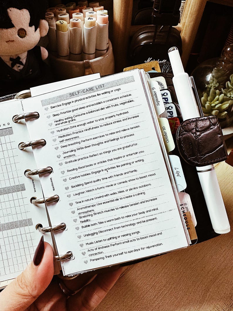 May include: A planner with a self-care list, including items like exercise, healthy eating, and meditation. The planner is open to a page with the title "SELF-CARE LIST". A white pen and a brown leather pen holder are also visible.