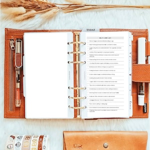 May include: A tan leather six-ring planner open to a self-care list page with gold hardware. The planner includes a clear pen holder, a white page with a checklist titled SELF-CARE LIST, and a matching tan leather pouch with a snap closure.